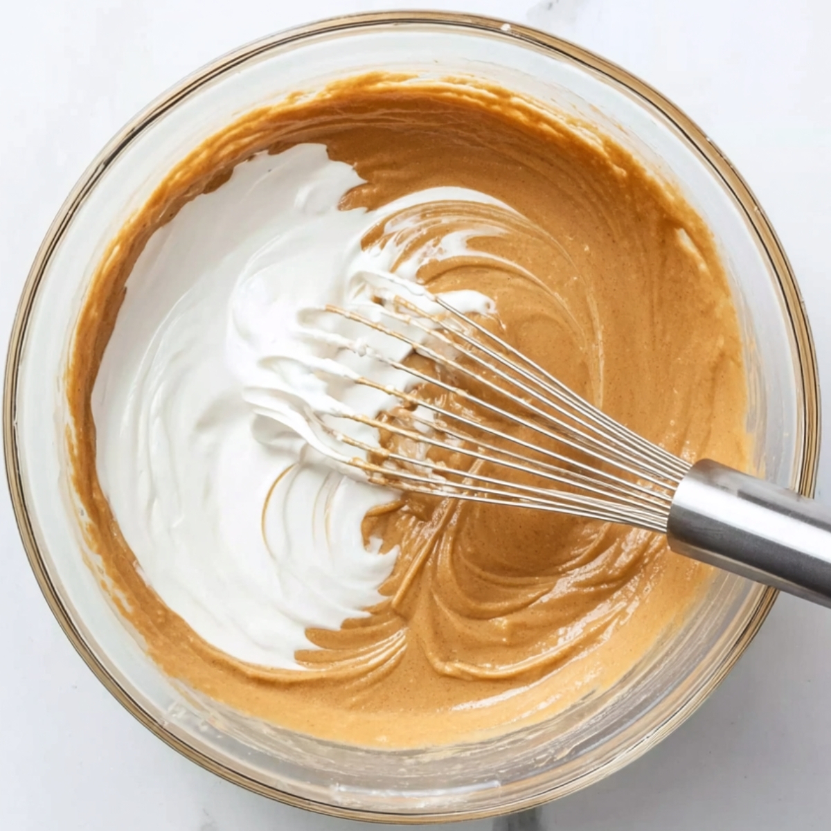 Overhead view of Biscoff batter in a glass bowl with white cream being whisked in using a metal whisk.