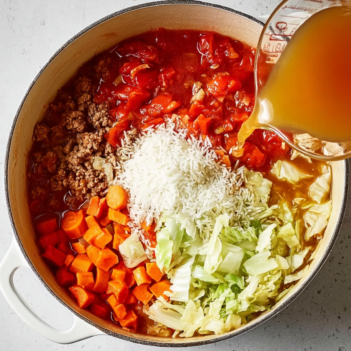 Top-down view of a white Dutch oven filled with raw cabbage roll soup ingredients including browned ground beef, diced carrots, chopped cabbage, uncooked white rice, and canned tomatoes, as broth is being poured in from a measuring cup.