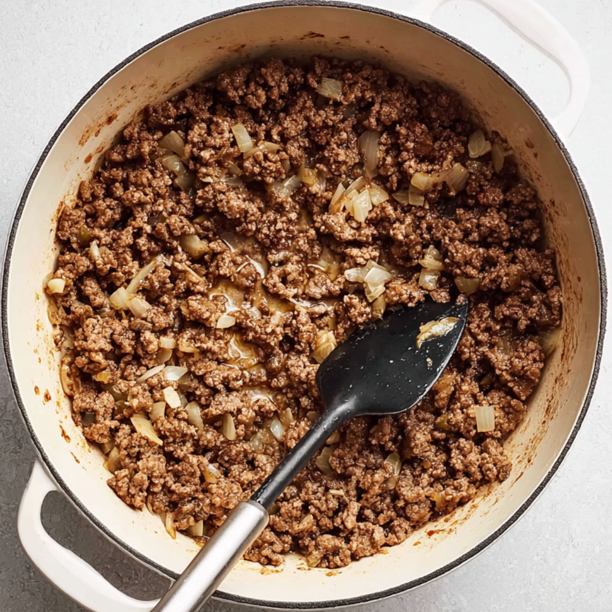 Overhead close-up of browned ground beef and diced onions cooking in a white enameled Dutch oven, with a black spatula resting inside the pot.