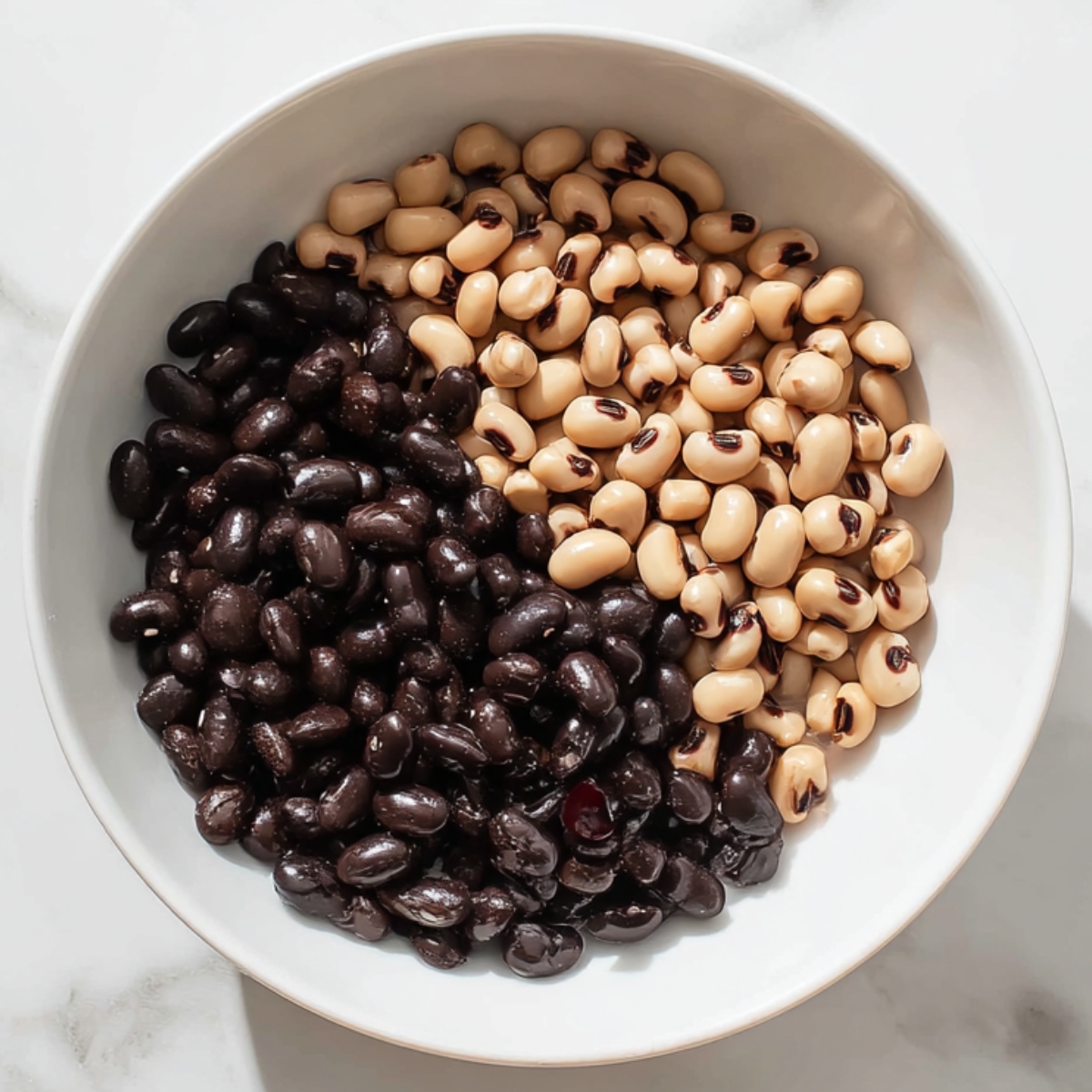 Top view of a white bowl with cooked black beans and black-eyed peas arranged side by side on a light marble countertop.