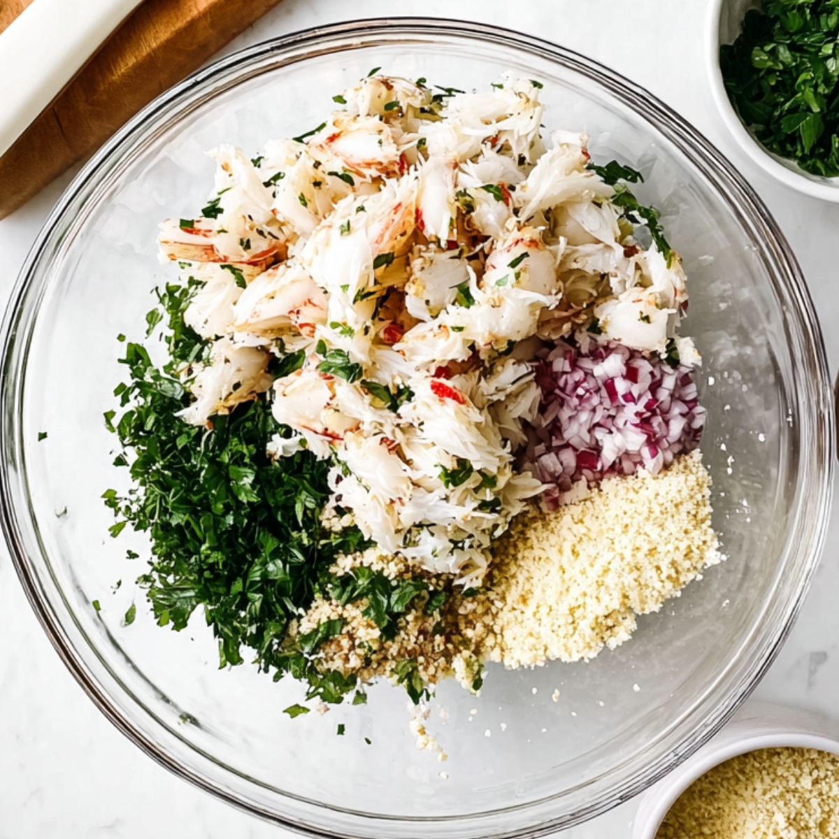 Glass mixing bowl filled with lump crab meat, chopped parsley, diced red onion, and breadcrumbs for homemade crab cakes on a marble countertop.