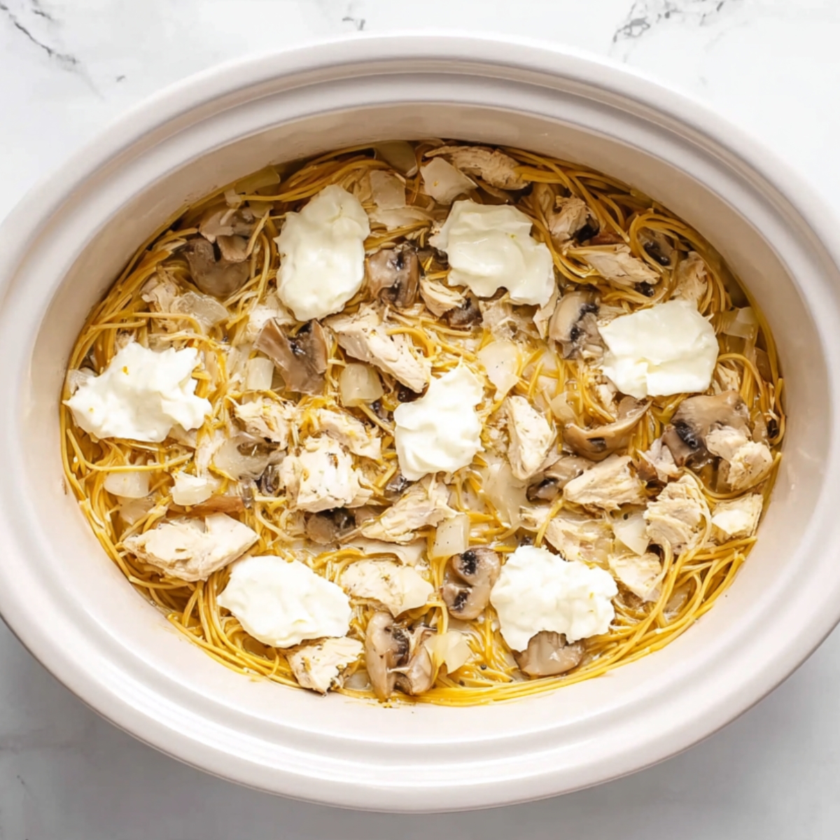 Crockpot Chicken Tetrazzini in slow cooker with spaghetti noodles, shredded chicken, sliced mushrooms, diced onions, and dollops of cream cheese before cooking.