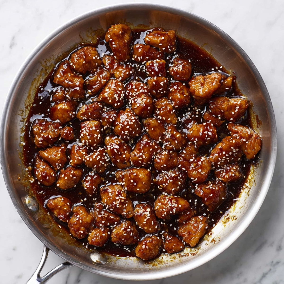 Succulent bite-sized sesame chicken pieces, glistening with a rich soy-based glaze, sizzle in a stainless steel skillet. The dark, caramelized sauce clings to the chicken, accented by a sprinkle of sesame seeds. Set against a clean white marble countertop, the dish is captured in vibrant, high-contrast lighting, showcasing its glossy, sticky texture in ultra-realistic detail.