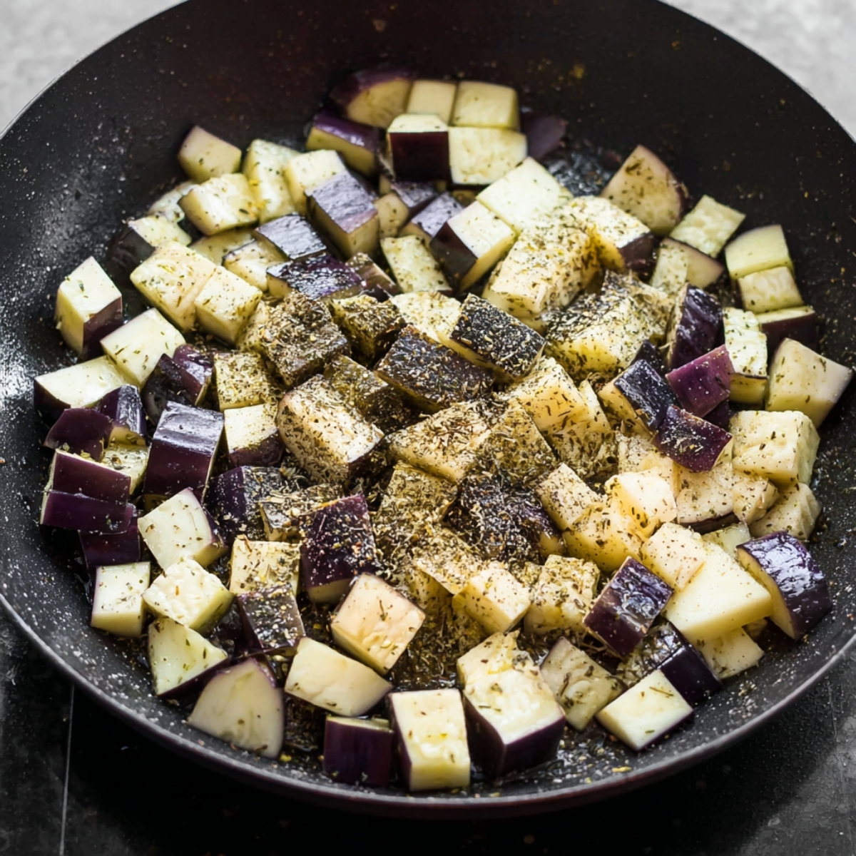 Close up of diced eggplant in a skillet, seasoned with oregano and black pepper, ready to be sautéed to perfection.