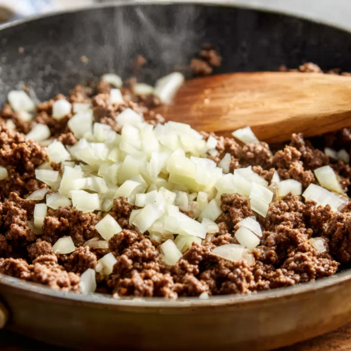 Ground beef cooking in a skillet with freshly diced onions being stirred with a wooden spoon, light steam rising from the pan.