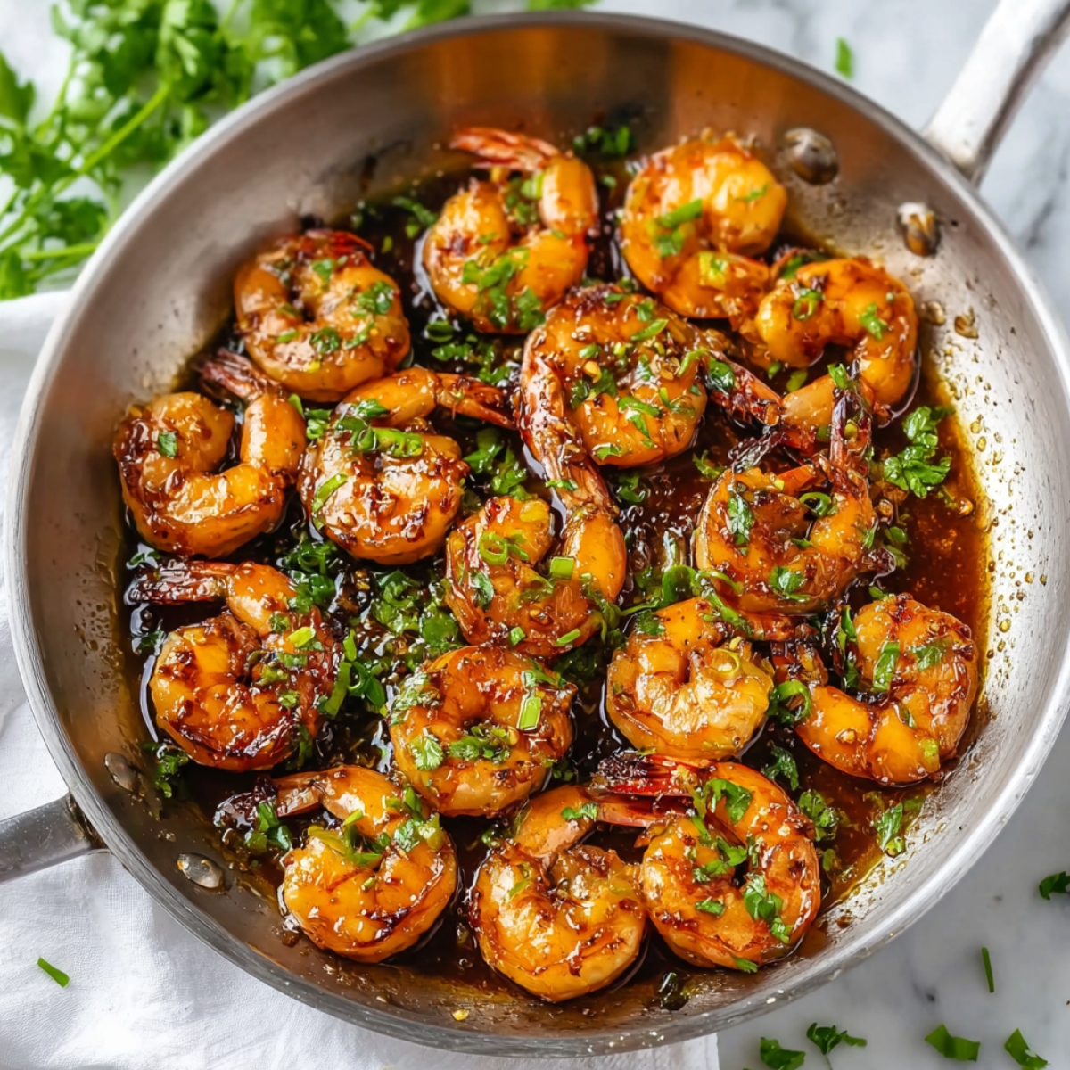 Honey garlic shrimp cooking in a stainless steel skillet, coated in a rich caramelized sauce and sprinkled with fresh herbs.