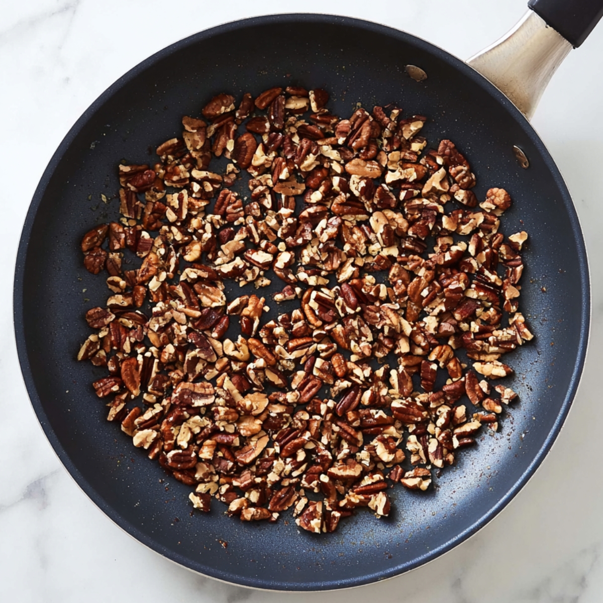 Overhead view of a black nonstick frying pan filled with finely chopped pecans being dry-toasted, nuts lightly browned and evenly scattered across the surface, set on a white marble countertop with subtle gray veining, soft natural daylight, minimal shadows, realistic food photography, high detail, sharp focus, clean modern kitchen aesthetic.