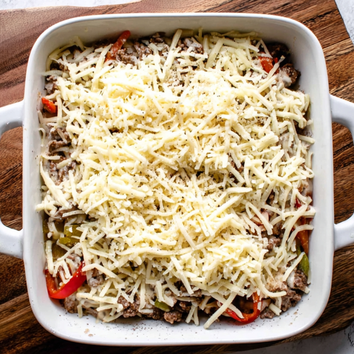 This image shows the casserole before it is fully baked, with shredded cheese sprinkled over the top. It shows the layers of seasoned beef and colorful peppers that will meld together once cooked.
