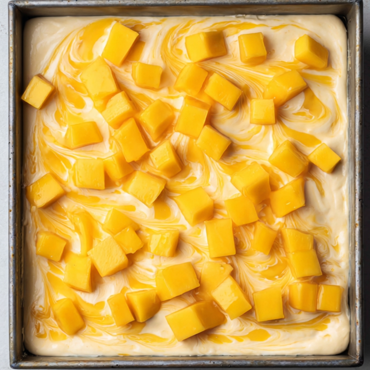 Top-down view of a square baking pan filled with creamy swirled mango mixture and evenly scattered fresh mango cubes on top, ready to chill or bake.