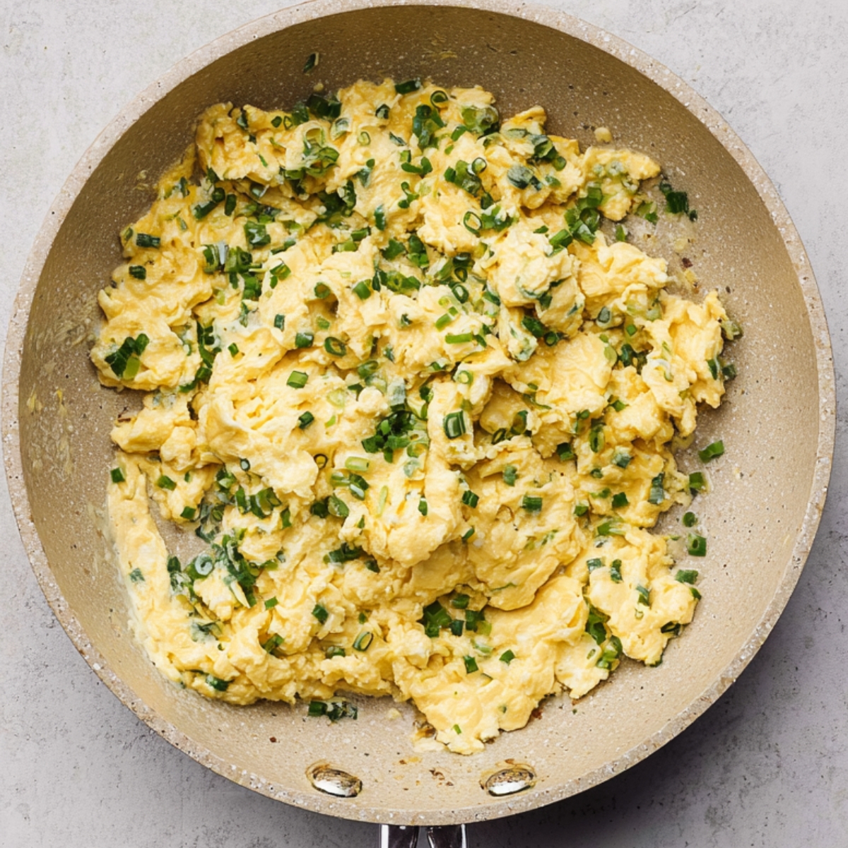 Top-down view of fluffy scrambled eggs mixed with chopped green onions cooking in a nonstick skillet on a light surface.