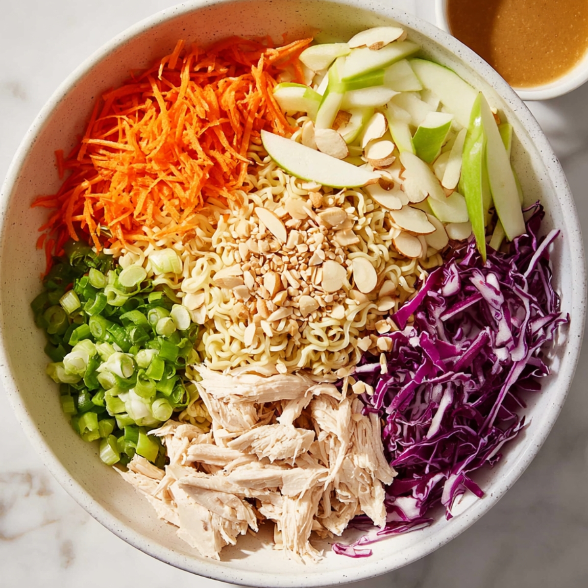 Fresh ramen noodle chicken salad ingredients beautifully arranged in a large white bowl. Crushed instant ramen noodles surrounded by shredded chicken, purple cabbage, carrots, green onions, sliced almonds, and julienned apple. A small bowl of sesame dressing on the side. Bright, clean, overhead food photography with natural lighting.