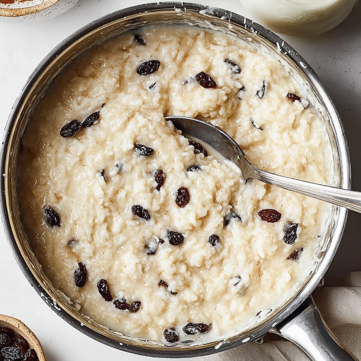Top view of thick rice pudding with raisins cooking in a saucepan, a spoon scooping from the creamy mixture.