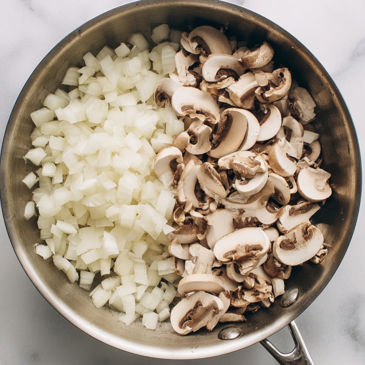 Top-down view of diced onions and sliced mushrooms in a stainless steel skillet on a marble countertop before cooking.