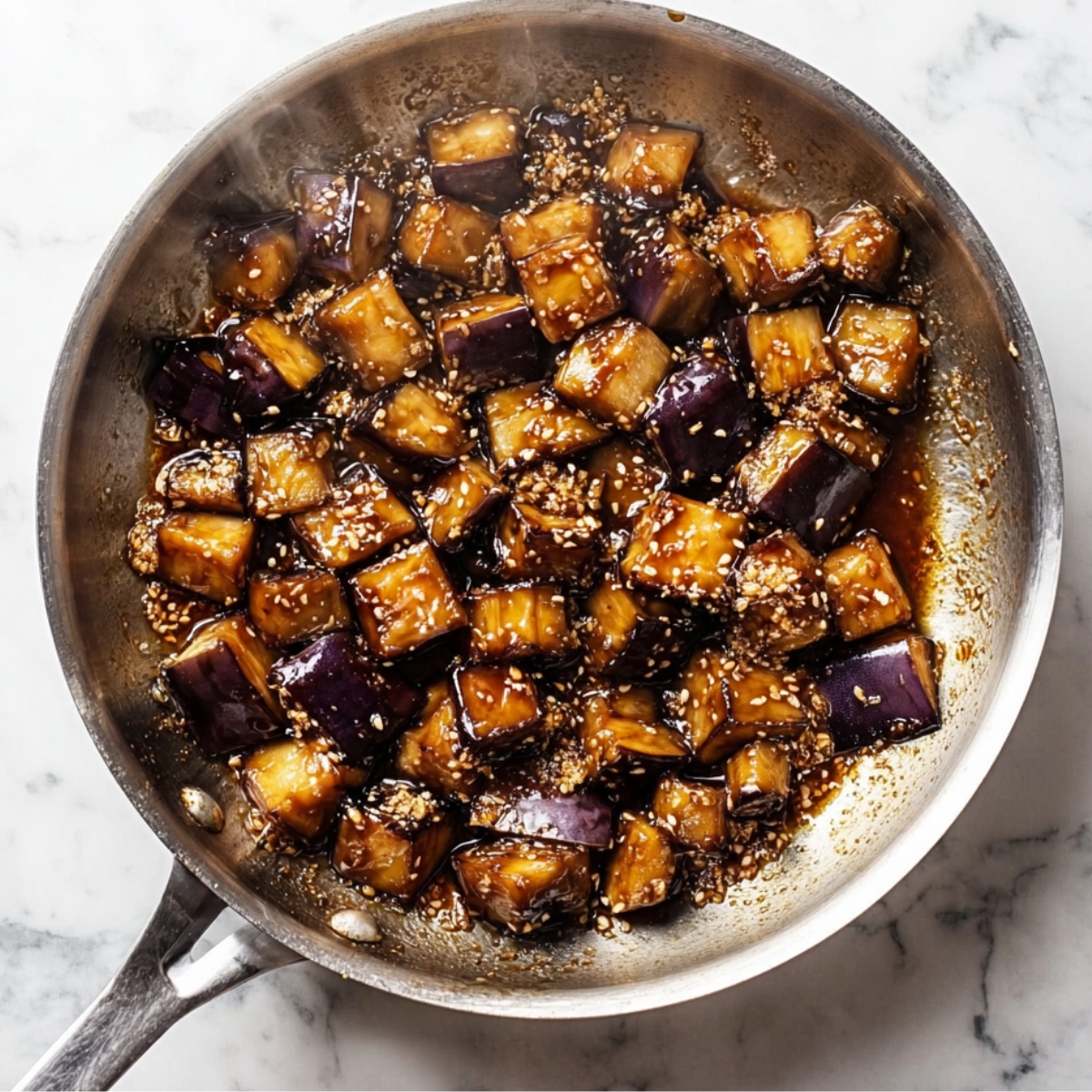 Overhead view of diced eggplant cooking in a skillet with thick soy glaze and sesame seeds, showing caramelized edges and rich brown sauce.