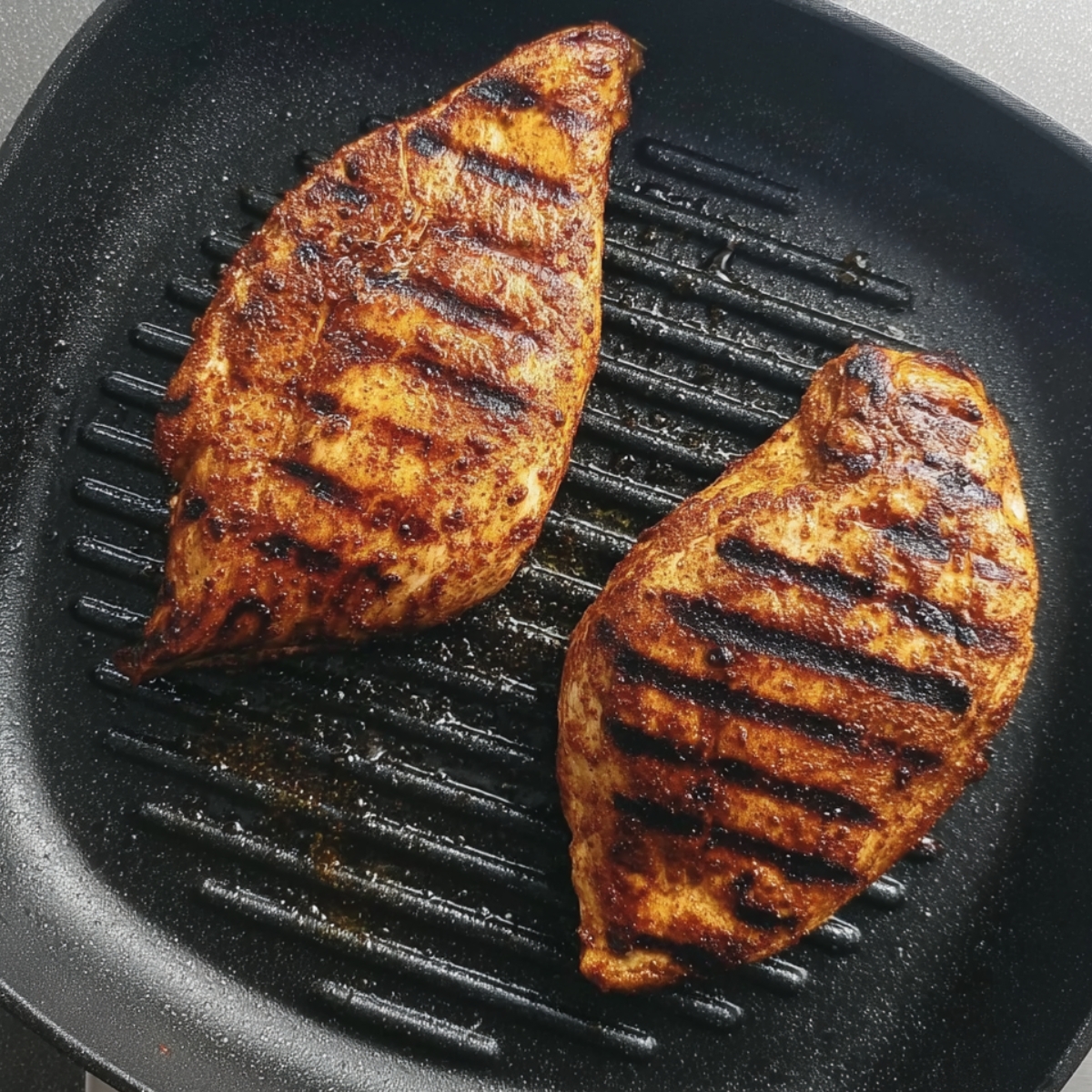 Two seasoned grilled chicken breasts cooking in a black ridged grill pan, golden brown with dark grill marks and slightly charred edges.