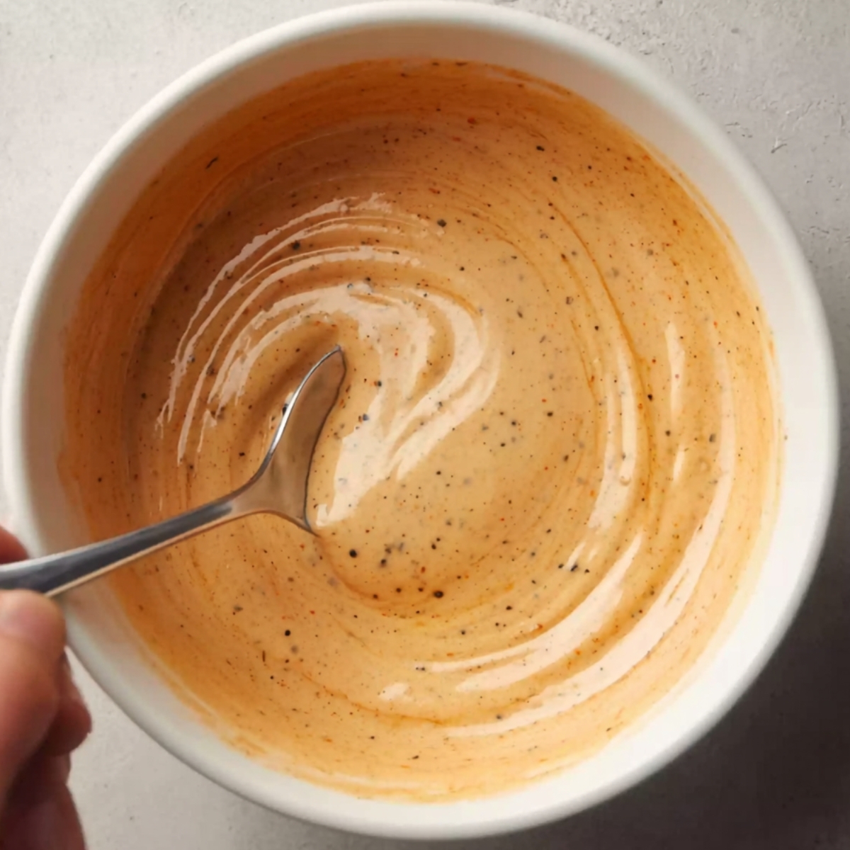 Top-down view of creamy orange chipotle southwest dressing being stirred with a spoon in a white bowl, with visible black pepper specks and smooth swirl texture on a light gray countertop.