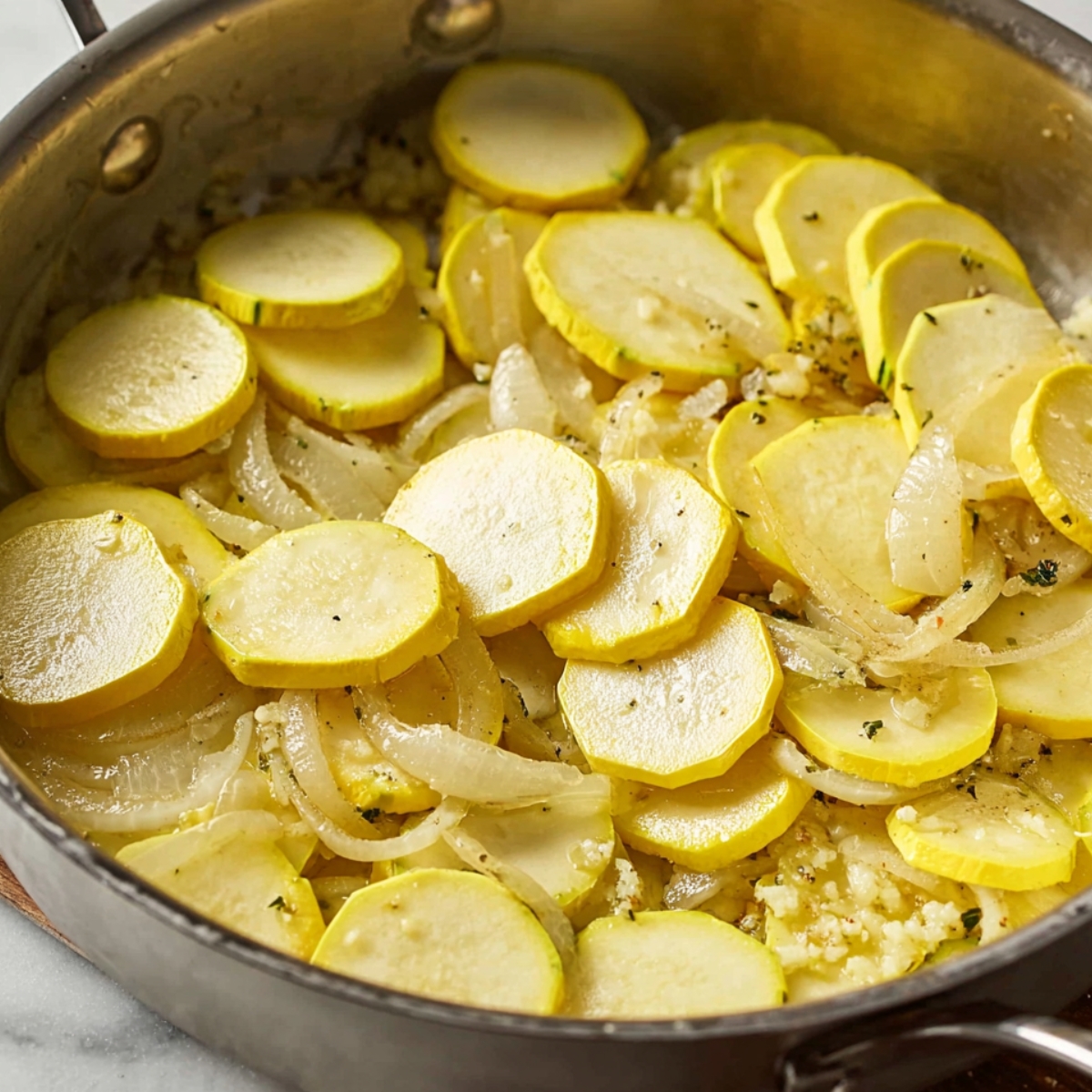 Close up of sautéed yellow squash and onions in a skillet, with garlic and fresh herbs adding flavor to the dish.