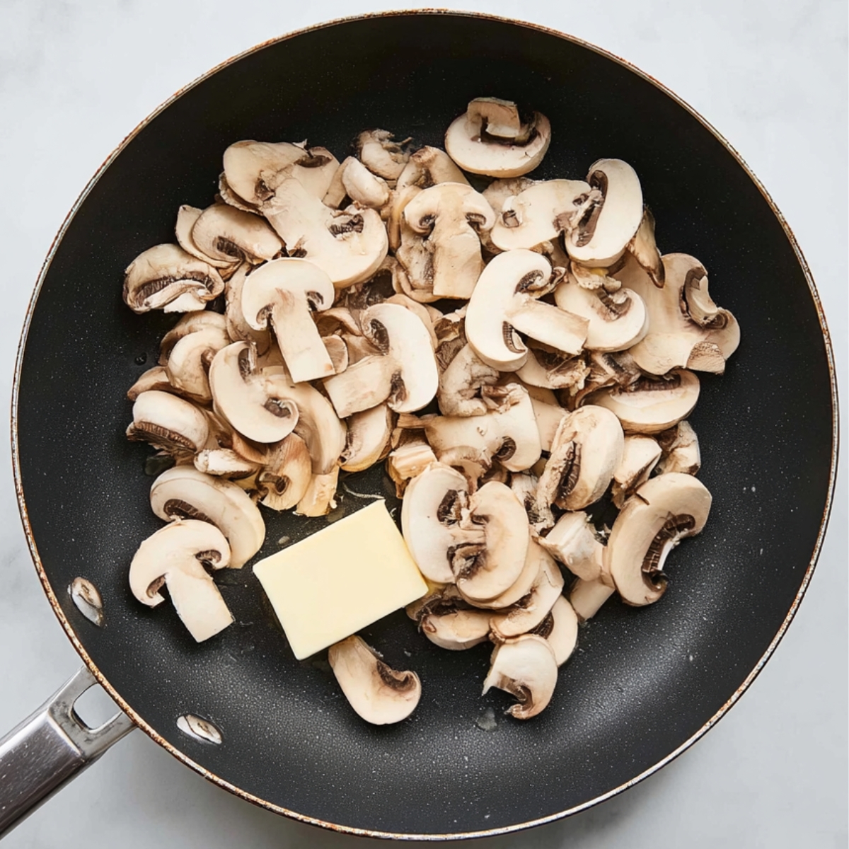 top-down view of a black nonstick frying pan on a white marble countertop filled with freshly sliced white mushrooms and a small cube of butter beginning to melt. Natural soft kitchen lighting, realistic textures, minimal composition, high-resolution food photography, neutral tones, sharp focus.