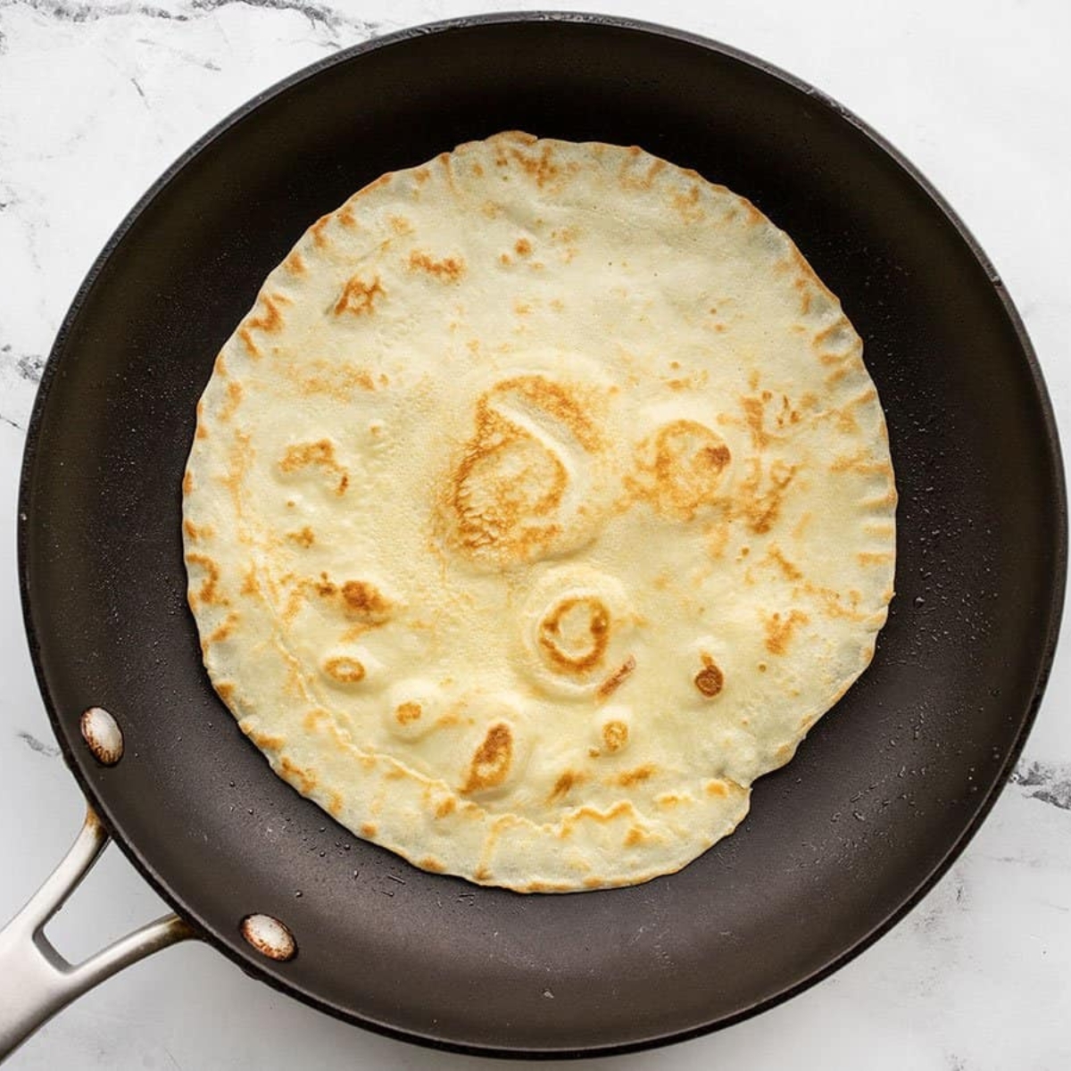 Overhead top-down food photography of a single flour tortilla cooking in a black nonstick frying pan. The tortilla has golden brown toasted spots and small air bubbles forming on the surface. The pan sits on a light marble countertop with a silver handle visible. Soft natural kitchen lighting, clean minimal background, realistic textures, high resolution, sharp focus, 4k detail.