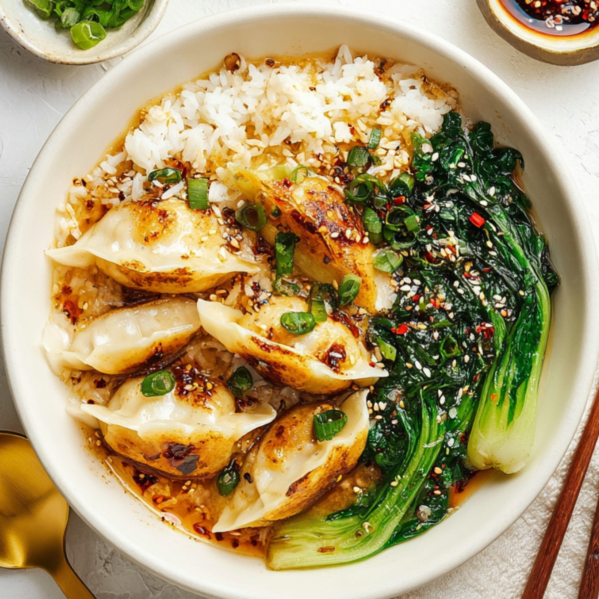 Brothy Rice with Ginger and Garlic Close-up image of a hearty dumpling bowl with pan-fried dumplings, steamed rice, and sautéed greens, topped with sesame seeds, chopped green onions, and spicy chili oil, emphasizing texture and color. The composition includes a gold spoon, black chopsticks, small condiment dishes, and a bowl of flavorful broth, all placed on a white textured surface to highlight the dish’s freshness, flavor, and visual appeal in a modern food photography style.