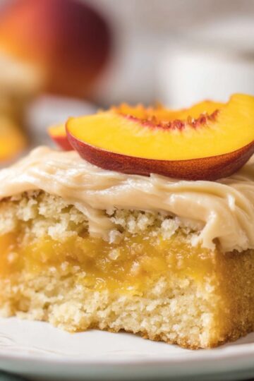 Brown Sugar Peach Cake Close up view of a slice of peach cake that looks freshly baked and ready to enjoy. The cake's tender yellow crumb is speckled with chunks of juicy peaches, while the thick frosting on top adds a creamy, indulgent layer of flavor. A peach wedge decorates the slice, enhancing the visual appeal and highlighting the fruitiness of the dessert. Shot with warm, natural light and a shallow depth of field, the image captures every texture detail-from the soft cake to the smooth frosting-making it look irresistible and perfect for food blogs, recipe posts, or social media content.