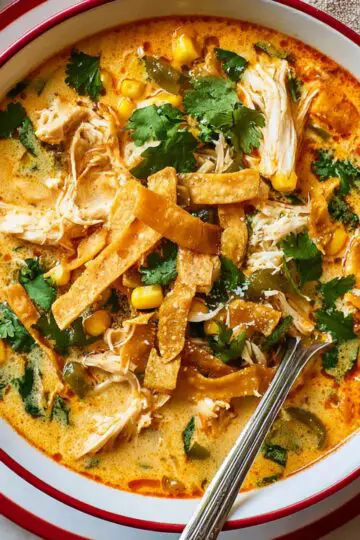Chicken Poblano Soup Close up of a creamy chicken poblano soup in a white bowl with red rim, topped with shredded chicken, crispy tortilla strips, sweet corn, and fresh cilantro, rich orange broth with a velvety texture, spoon resting in the bowl, overhead food photography, vibrant colors, natural lighting, cozy homemade comfort food scene, high detail and appetizing presentation