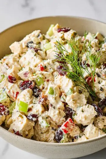 Chicken Salad with Fresh Apples Close up of a creamy chicken salad in a beige bowl, filled with diced chicken, celery, red bell peppers, dried cranberries, and herbs, lightly coated in a rich dressing, garnished with fresh green sprigs, soft natural lighting, marble countertop background, high detail, food photography, shallow depth of field, realistic texture, appetizing presentation