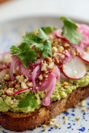Cilantro Lime Toast Close up of a slice of toast topped with creamy green spread, pickled red onions, fresh cilantro, radish slices, and crushed nuts, served on a white plate with blue speckles, natural lighting, high detail, shallow depth of field, gourmet food styling.