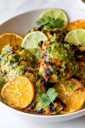 Easy Mojo Chicken Close up of juicy grilled chicken pieces coated in vibrant green cilantro sauce, garnished with fresh lime slices, orange slices, and scattered cilantro leaves, served in a white bowl on a light marble surface, natural lighting, high detail, food photography, fresh and healthy aesthetic, shallow depth of field, bright and clean composition, appetizing and colorful plating