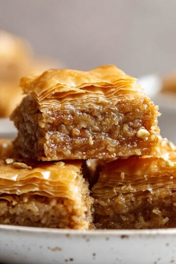 Greek Baklava recipe Close up of golden homemade baklava squares stacked on a white ceramic plate, showing delicate layers of flaky phyllo pastry and finely chopped nut filling soaked in syrup, warm natural lighting, soft blurred kitchen background, rich textures and crispy edges visible, Mediterranean dessert presentation, high-resolution food photography with shallow depth of field.