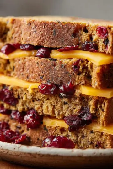 Jamaican Spice Bun Close up of stacked slices of homemade spiced bread filled with cranberries, each layer separated with a slice of cheddar cheese. The photo showcases the crumbly yet soft texture of the bread, rich golden-brown crust, and vibrant red berries, creating a cozy and appetizing presentation. Taken with shallow depth of field, the background is blurred, emphasizing the bread's details and textures. Perfect for highlighting breakfast or snack ideas, festive holiday recipes, or creative cheese and bread pairings. Ideal for Pinterest boards, food blogs, or social media posts targeting home bakers and culinary enthusiasts looking for easy, flavorful, and visually stunning baked goods.