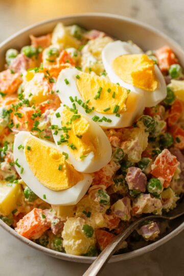 Olivier Potato Salad Close up of a bowl of Russian Olivier salad with diced carrots, potatoes, peas, ham, chopped boiled eggs, and green onions, topped with three slices of boiled egg and garnished with chopped chives, served with a fork on a marble countertop, warm natural lighting, high detail, vibrant and appetizing food photography style