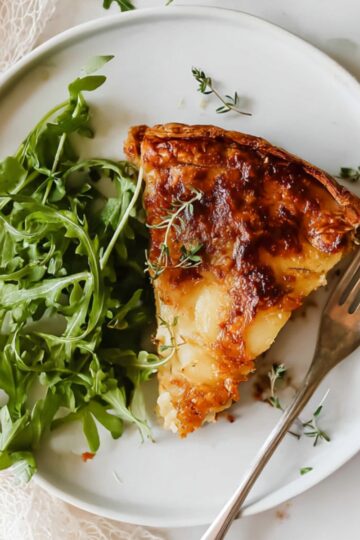 Potato Galette Close up of a slice of cheesy potato galette on a white plate, garnished with fresh arugula, fork resting on the side, natural light highlighting the golden-brown crust, overhead view, styled food photography, minimalistic background with soft textures, cozy and inviting atmosphere.