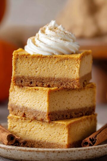 Pumpkin Cheesecake Bars Close up of three stacked pumpkin cheesecake bars with a golden crust, topped with a swirl of whipped cream, set on a ceramic plate with cinnamon sticks nearby, blurred pumpkins in the background, warm natural lighting highlighting the creamy texture and autumnal colors, high-resolution food photography.