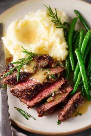 Steak with Garlic Cream Sauce Close up of a plated steak dinner with sliced medium-rare steak topped with creamy garlic sauce, fresh green beans, and buttery mashed potatoes, garnished with rosemary, fork and knife beside plate, natural lighting, high detail, appetizing food photography style