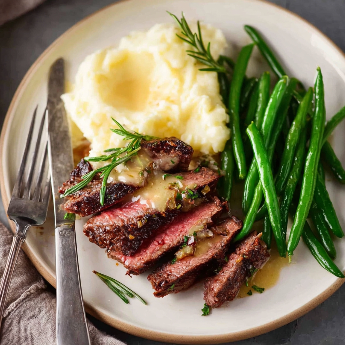 Steak with Garlic Cream Sauce Close up of a plated steak dinner with sliced medium-rare steak topped with creamy garlic sauce, fresh green beans, and buttery mashed potatoes, garnished with rosemary, fork and knife beside plate, natural lighting, high detail, appetizing food photography style