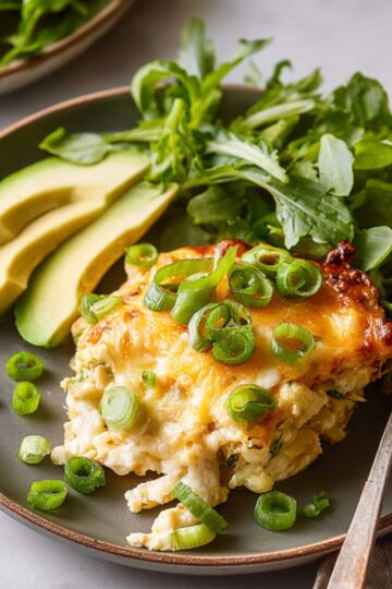 Tater Tot Breakfast Casserole Close up of a creamy baked casserole topped with melted cheese and fresh green onions, served on a modern plate with sliced avocado and a side of leafy greens, soft natural lighting, high detail food photography, warm tones, minimal background, rustic kitchen aesthetic, shallow depth of field, appetizing texture, overhead composition, Instagram-worthy plating
