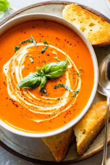 Tomato Bisque Soup Close up of a creamy tomato bisque in a white bowl, garnished with fresh basil and a swirl of cream, served with toasted bread on a rustic plate, bright natural lighting, overhead view, vibrant colors, food photography style