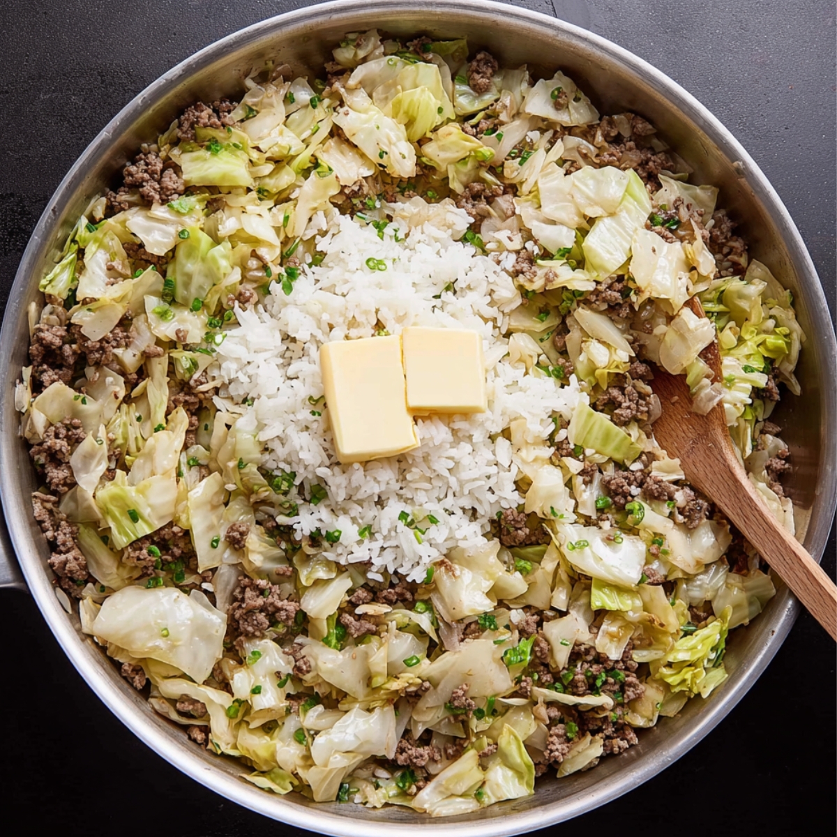 Close up of a skillet with rice, cabbage, and beef, topped with butter – a comforting meal coming together.