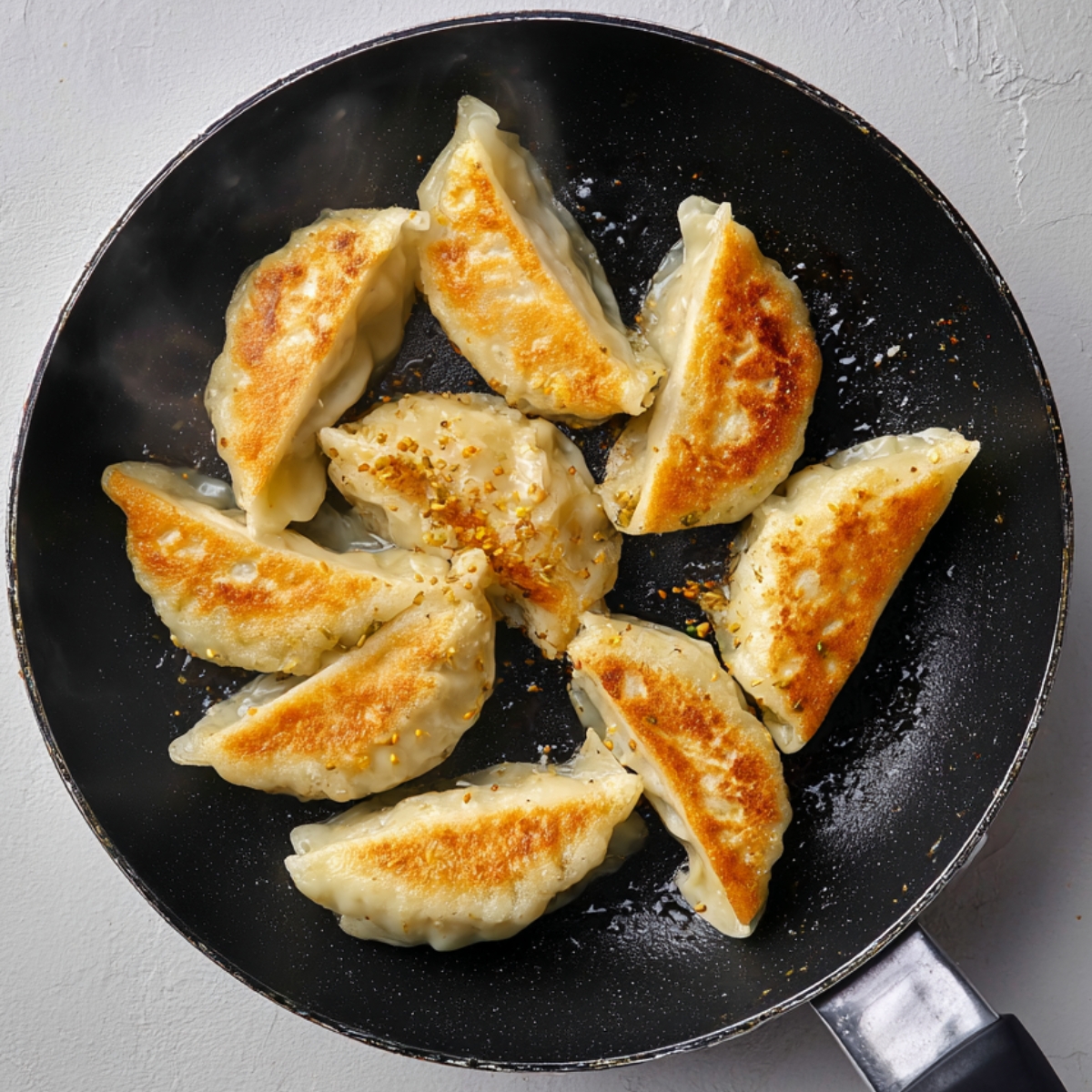 A close-up top-down view of a delicious bowl of pan-fried dumplings served with fluffy white rice and vibrant sautéed greens, drizzled with spicy chili oil and sprinkled with sesame seeds and fresh chopped scallions. The scene includes a gold spoon, black chopsticks, small condiment dishes, and a steaming bowl of broth on a clean white textured background, showcasing a beautifully arranged, colorful, and appetizing Asian-inspired meal.