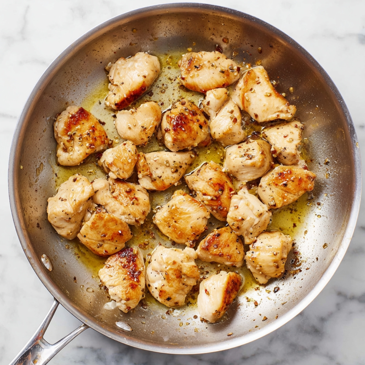 Close up of golden-brown cooked chicken pieces in a stainless steel frying pan, with slight browning and oil glistening, on a white marble countertop, natural lighting, high detail texture, home cooking scene.