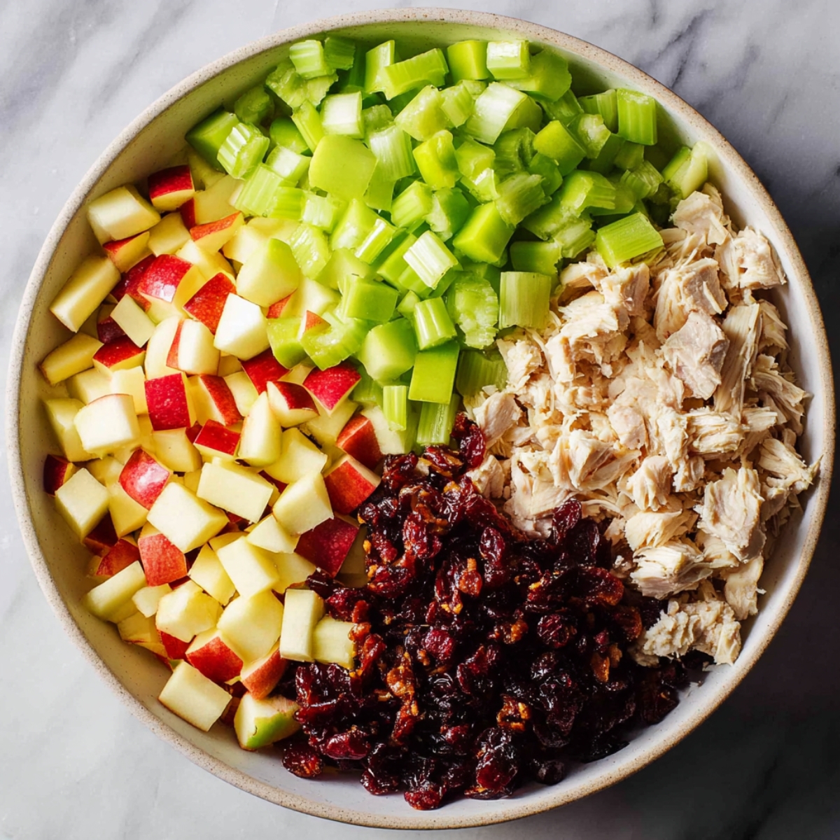 Close up of a large bowl filled with neatly arranged chopped ingredients, including diced green celery, cubed red-skinned apples, shredded cooked chicken, and a portion of dark dried cranberries, all separated into sections in a light ceramic bowl on a marble surface, soft natural lighting, vibrant colors, high detail, food photography style.