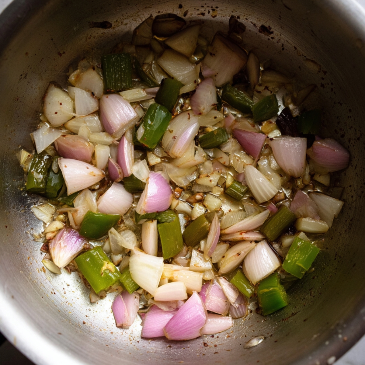 Close up of sautéing chopped garlic, shallots, and jalapeños in a stainless steel pan, showing sizzling vegetables with golden-brown edges, natural lighting, high detail, overhead angle.