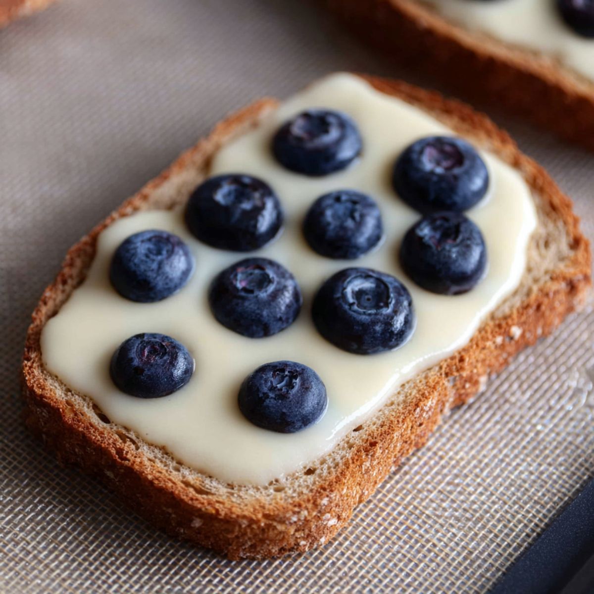 Close up of a slice of whole wheat bread topped with creamy custard and fresh blueberries, placed on a silicone baking mat, natural lighting highlighting the texture and colors, minimalistic food photography style, high detail, soft shadows.