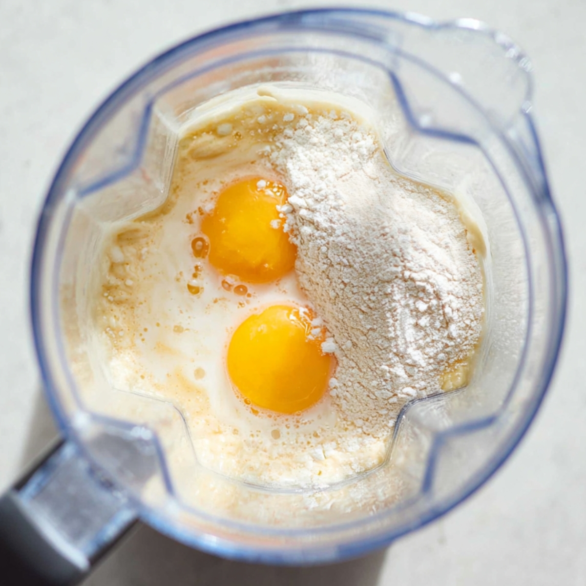 Close up of a blender filled with pancake batter, showing raw eggs, flour, and milk ready to be mixed, bright natural lighting, top-down view, detailed texture of ingredients.