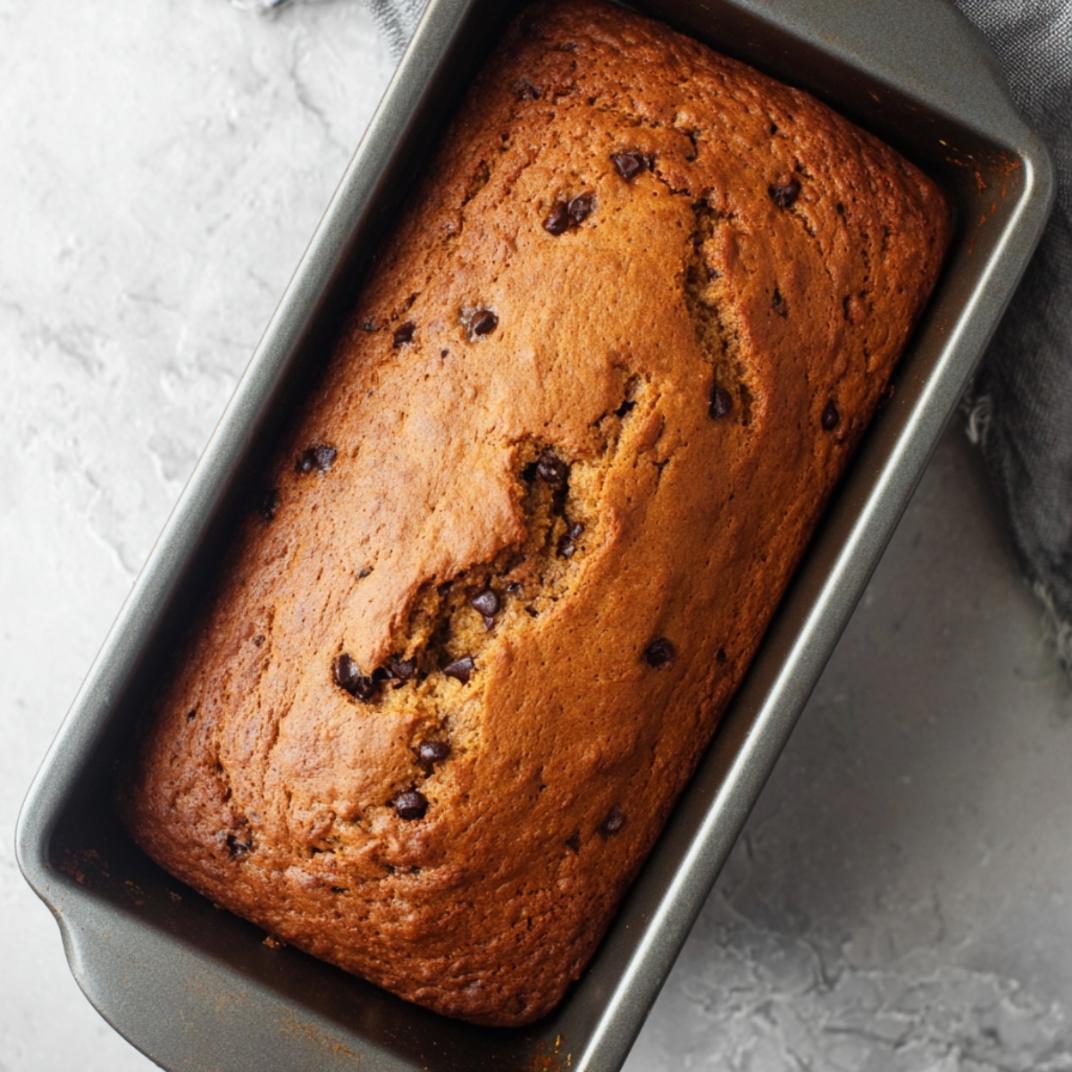 Close up of a freshly baked brown loaf in a rectangular baking pan, showing a golden-brown crust with slight cracks along the top. The loaf is rich and moist, with subtle chocolate chips or fruit pieces peeking through the surface, inviting you to slice and enjoy. This overhead view highlights the perfectly risen texture and realistic crumb, ideal for recipes like banana bread, pumpkin loaf, or fruit bread. Perfect for food photography, baking guides, or recipe posts that inspire home bakers to create their own delicious homemade breads. The natural lighting and clean kitchen countertop background make the loaf visually appealing and ready for Pinterest sharing.