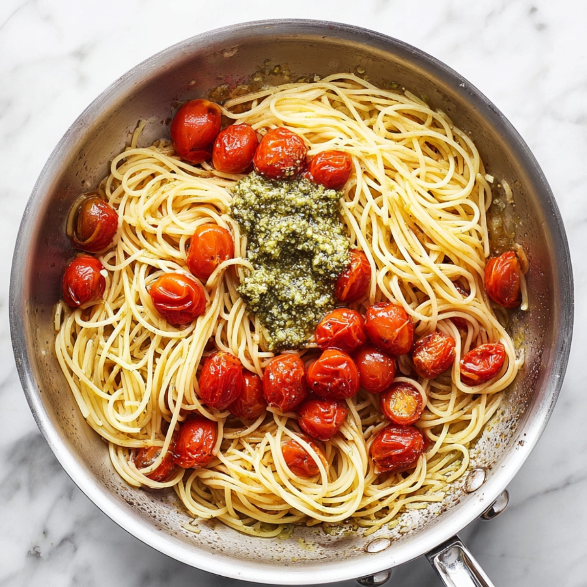 Close up of cooked spaghetti with cherry tomatoes and a dollop of green pesto in a stainless steel frying pan on a white marble countertop, highlighting the textures and colors of the pasta and sauce, top-down view, natural lighting, food photography, appetizing presentation.