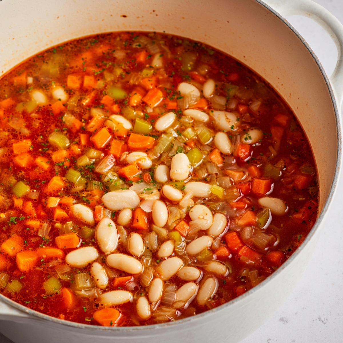 Close up of a bowl of creamy vegetable and bean soup, featuring a rich orange broth filled with tender carrots, celery, and white beans, garnished with fresh green parsley and a wedge of lemon. The white ceramic bowl is placed on a simple light-colored surface, with a silver spoon beside it and parsley leaves casually scattered around. The lighting is soft and diffused, creating bright highlights on the creamy surface and emphasizing the fresh, colorful ingredients. The composition is clean, minimalistic, and visually appealing, ideal for a modern food photography style.
