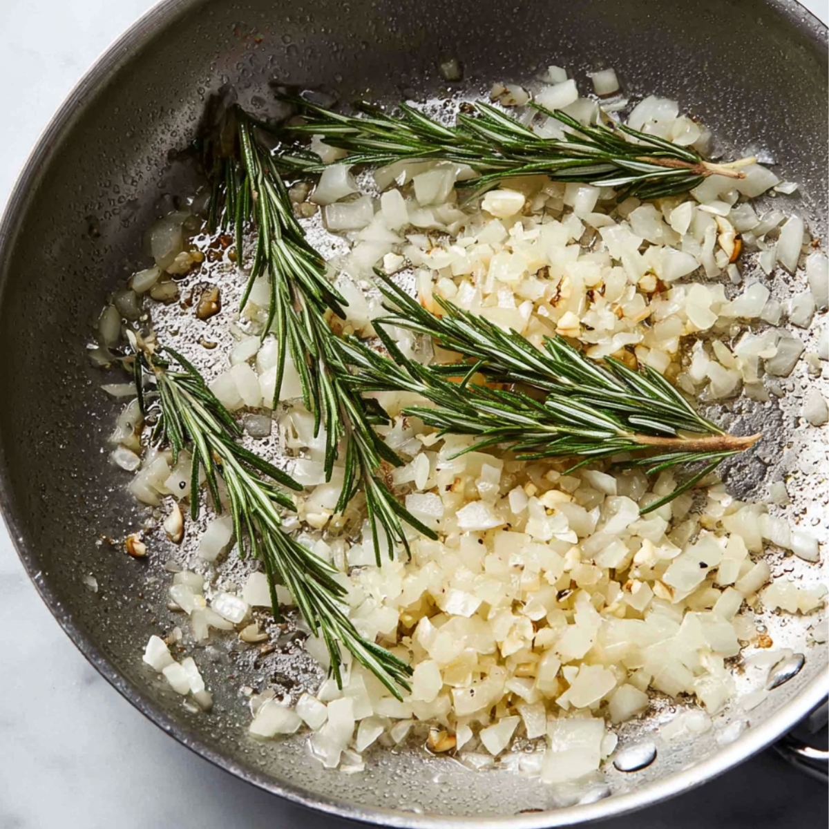 Close up of sautéed onions, garlic, and rosemary in a pan, with the ingredients gently cooking together to release aromatic flavors.
